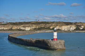 The entrance to Newhaven harbor, East Sussex, UK, and its breakwater and lighthouse. Clear blue sky and water with white cliffs in the background.