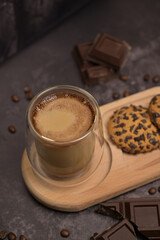 Close up. Coffee in a transparent glass. Oatmeal cookies on a wooden board on the table. Pieces of broken dark chocolate. Spilled coffee beans. Dark background. Coffee service. Still life