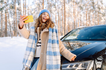 A beautiful woman with phone takes selfie in snowy forest next to car. Young traveler blogs and...