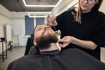 Woman Barber shaving bearded customer in modern style barbershop. Male Beard hair cutting with scissors. Man client visit gentleman grooming beauty salon for evens the beard