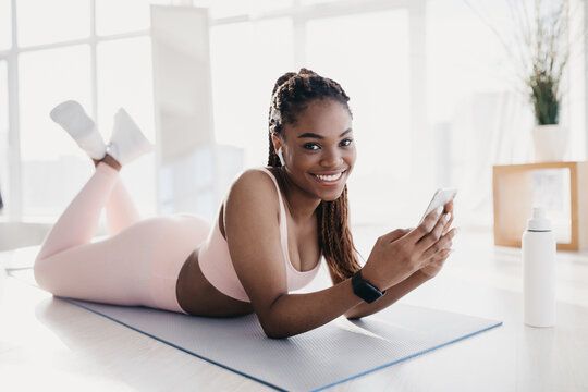 Smiling young woman lies on a yoga mat at home, taking a break from her workout. She listens to her favorite music through earphones while using her smartphone.