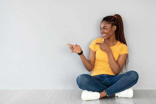 A happy young black woman sits cross legged on a grey studio floor. She is playfully pointing to the side, inviting viewers to consider the empty space for promotional content.