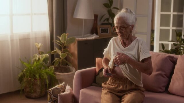 Medium full shot of gray-haired old Caucasian woman giving herself insulin injection in abdomen, lifting up blouse, while controlling diabetes. Glucose test kit lying on table in cozy living room