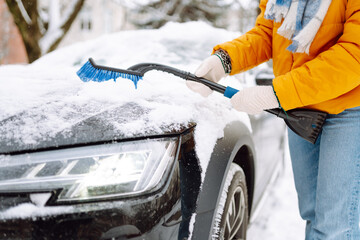 Happy woman in bright jacket and hat clears snow from her car on winter morning. Close-up of a...