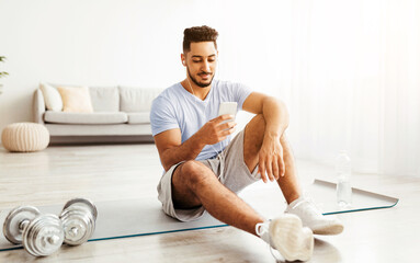 Young man sits on exercise mat in a well-lit room, taking a moment to check his phone after a workout. Dumbbells are placed nearby, showing a commitment to fitness.