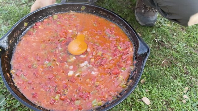 Cracking eggs into a delicious menemen or sausage and egg dish prepared in an open-air cast iron pan.