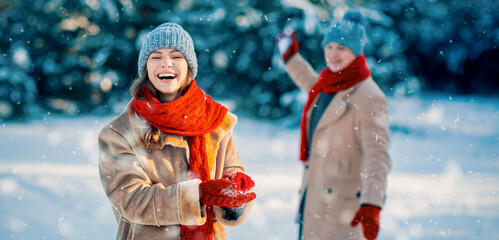 Friends are having a fun day in the snow. One person smiles and holds snow in hands while the other waves. Both wear warm clothing, surrounded by softly falling snowflakes.