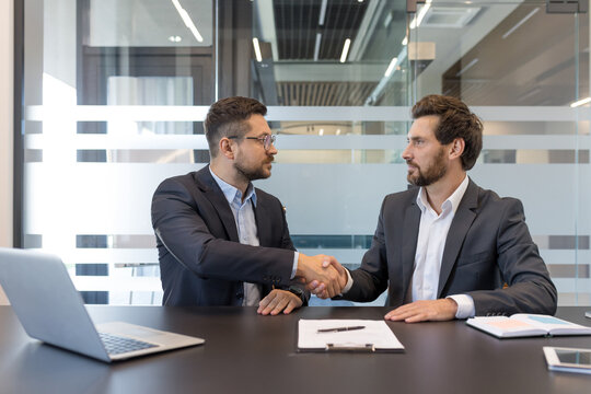 Two businessmen shaking hands across a conference table after closing a deal, smiling partners finalizing a contract in a modern office meeting, signaling cooperation and success - Powered by Adobe