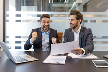 Two excited businessmen celebrating business success, looking at a chart with rising profits, demonstrating teamwork and achieving financial growth in an office meeting