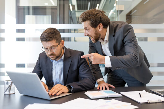 Angry boss pointing and yelling at frustrated male employee over laptop in a tense office scene, depicting workplace bullying, stress, pressure, conflict and discrimination
