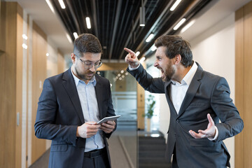 Two businessmen having an argument in an office hallway, with a stressed employee looking down at a tablet while his angry boss points and yells, representing workplace conflict and harassment