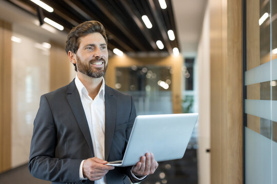 Businessman in a suit and white shirt holding a laptop and smiling, looking away with a thoughtful expression in a contemporary corporate building corridor - Powered by Adobe