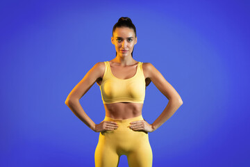 A fit model stands with hands on hips, showcasing a yellow sports outfit. The backdrop is a vibrant blue, creating an energetic atmosphere. This scene emphasizes fitness and confidence.