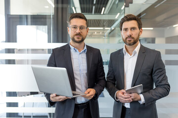 Two professional businessmen standing in a contemporary office building, one holding a laptop and the other a digital tablet, representing partnership and confident teamwork
