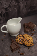 Milk jug and oatmeal cookies on the table. Pieces of broken dark chocolate. Spilled coffee beans. Dark background. Coffee service. Still life