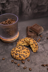 Coffee beans in a transparent glass, oatmeal cookies on the table. Pieces of broken and oatmeal cookies on the table. Pieces of broken dark chocolate. Spilled coffee beans. Dark background. Still life
