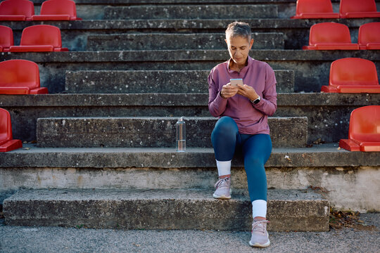 Mature athletic woman using smart phone while relaxing on staircase at stadium.
