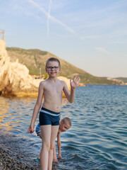 Two boys in swimwear on rocky beach at sunset, one waving and smiling to camera. Concept of summer vacation, childhood joy, travel freedom.