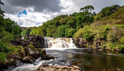 Fototapeta premium Cascading Waterfall Over Dark Rocks Surrounded by Lush Greenery and a Cloudy Sky during Daytime