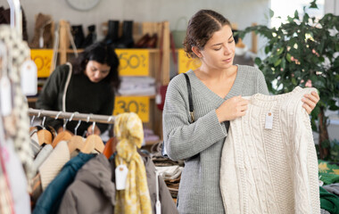 European woman chooses a knitted jersey sweater against the background of a rail and a rack with clothes in a store. Customer makes bargain purchases in a second hand. Woman supports slow fashion