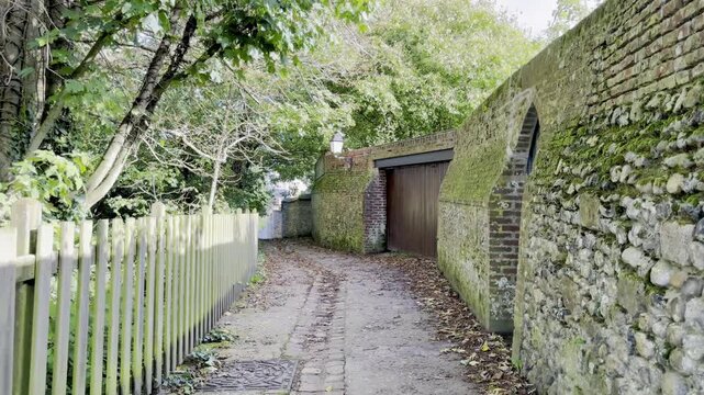 Rue du Beffroi in Saint-Valery-sur-Somme, narrow leafy passage with flint rampart walls, lanterns and cobbled wheel ruts leading between gardens and brick houses in the medieval upper town