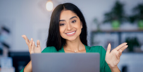 A woman with long black hair smiles while speaking animatedly at her laptop in a bright workspace filled with plants. She appears friendly and engaged in a virtual discussion.