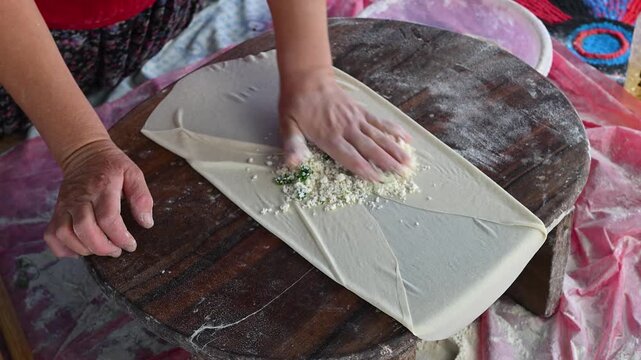 A woman adding cheese and parsley filling to yufka dough on a traditional wooden table and folding it into g&ouml;zleme.
