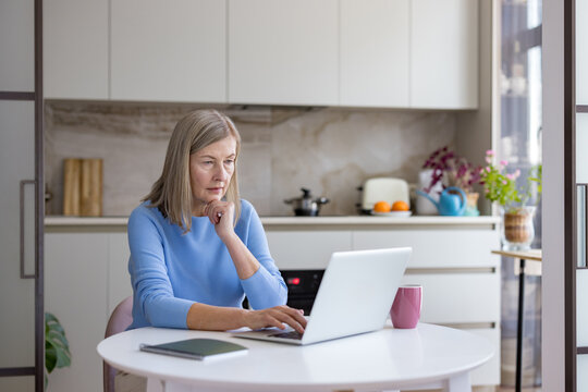 Senior businesswoman concentrating while typing on a laptop computer, working remotely from her modern kitchen table, showcasing digital literacy and an active lifestyle