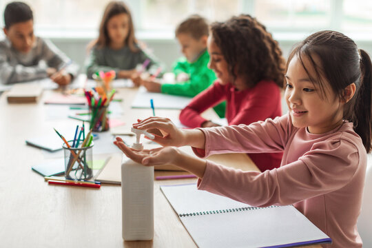 In a bright classroom, a schoolgirl uses hand sanitizer as she sits with her classmates, ensuring health safety during lessons. They focus on their work amidst ongoing pandemic precautions. - Powered by Adobe