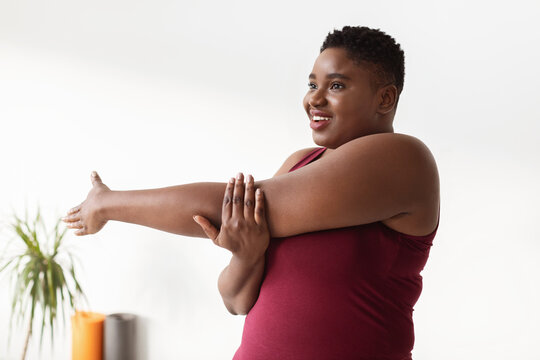 A smiling plus size millennial woman stretches her arms in a yoga studio, standing beside a white wall, promoting body positivity and fitness in a calm environment.