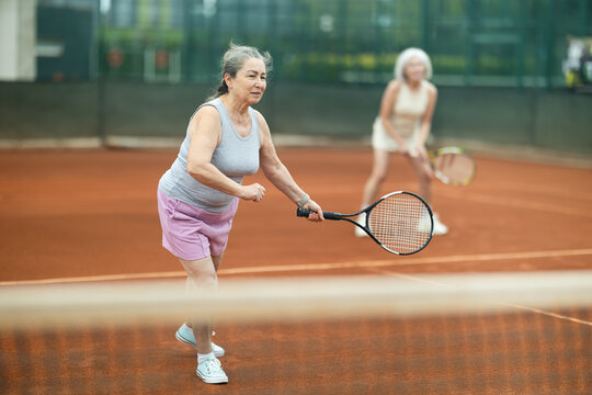 Focused positive elderly woman engrossed in tennis game on clay court, playing doubles with female partner captured in background, showcasing fitness and teamwork - Powered by Adobe