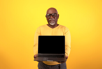 Senior black man smiles while holding a laptop with a blank screen in front of an orange studio background, promoting a website concept related to technology and internet use.
