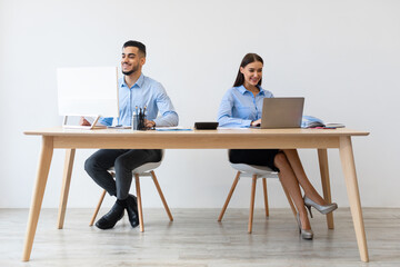 Diverse colleagues work together in a modern office setting, both using laptops at their desks. They are smiling and appear to enjoy their collaborative environment.