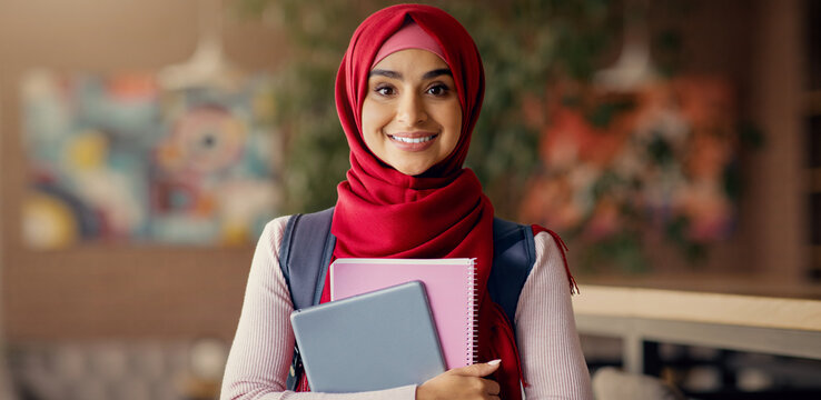 A young student with a red hijab stands confidently, holding a stack of books and a tablet. She is inside a cozy, modern space with art on the walls, smiling warmly. - Powered by Adobe