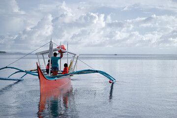 Obraz premium A family enjoys a peaceful day on a colorful red and blue outrigger boat, gliding over tranquil water as white clouds drift above, capturing togetherness and adventure at sea.