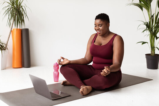 Young plus size black woman wearing sportswear enjoys yoga at home. She sits in lotus position on a fitness mat, smiling while following an online class on her laptop. - Powered by Adobe