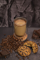 Coffee cappuccino latte in a transparent glass, oatmeal cookies on the table. Pieces of broken dark chocolate. Spilled coffee beans. Dark background. Coffee service. Still life