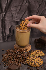Close up. Woman's hand dips a piece of oatmeal cookie into coffee cappuccino latte in a transparent glass on the table. Pieces of broken dark chocolate. Spilled coffee beans. Dark background