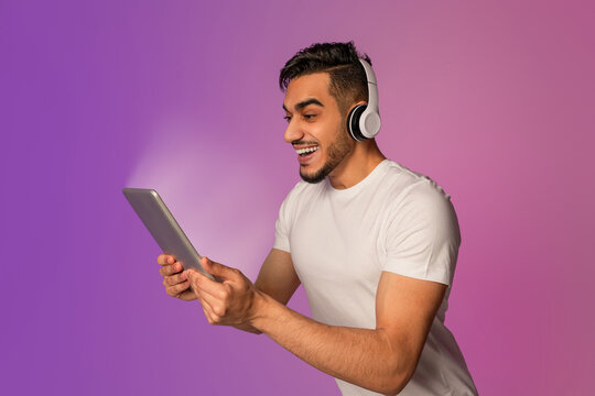A young Arab guy is excitedly looking at a glowing tablet, wearing headphones. He is overjoyed about a huge sale while shopping online. The scene is illuminated with neon colors. - Powered by Adobe