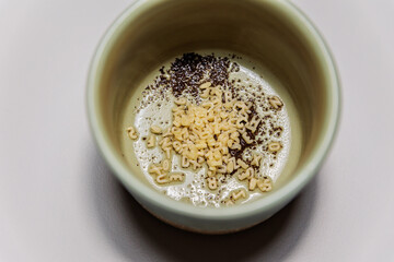 Close-up of a ceramic cup containing a mix of small alphabet-shaped pasta pieces and coffee grounds on a foamy surface, creating an abstract and artistic food composition