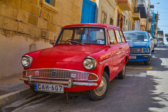 Close front view of red vintage Ford wagon parked by stone buildings and blue doors: Xghajra, Malta - June 7, 2021