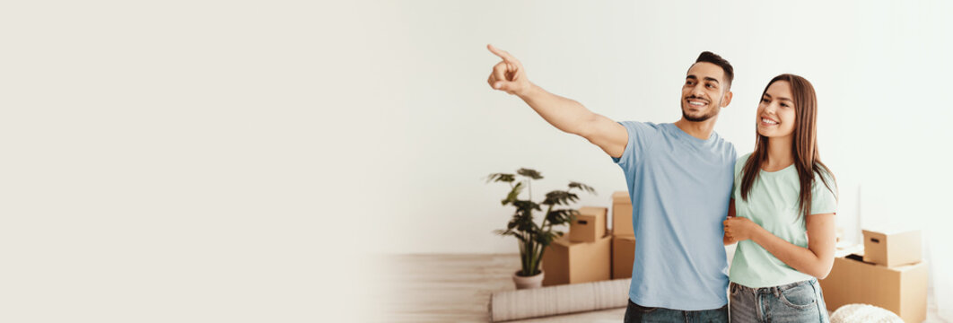 A happy couple stands in their new living space, surrounded by moving boxes. They are smiling and pointing, excitedly discussing their future plans for the home.