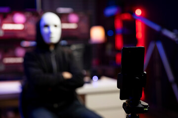 Close up of masked man using phone to film video, doing espionage campaign from hidden bunker. Focus on smartphone used by scammer wearing anonymous mask to record demands from secret location