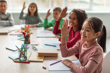 Multiethnic school kids sit at their desks in a bright classroom, eagerly raising their hands to answer questions. They smile, showing excitement and enthusiasm for learning.