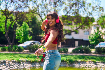 A beautiful Latina woman dances with rhythm and agility. She wears pink headphones, makeup and jeans. She is dancing in an outdoor park in a retro style. She exudes attitude, style, freedom and beauty
