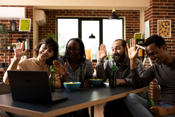 Four people concentrating on virtual call on laptop, making hello gesture. Diverse group of adults sitting with drinks during videoconference with long distance friends in relaxed apartment setting.