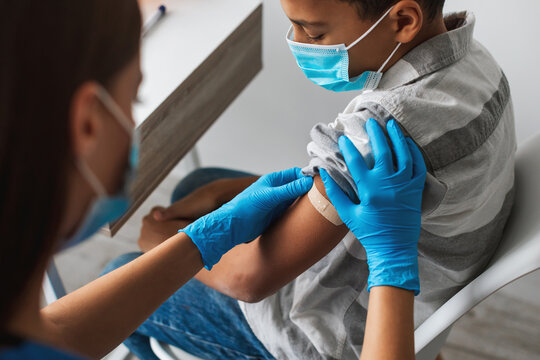 A young African American boy sits indoors wearing a face mask as a medical worker administers a vaccine. The child is ready to receive protection against Covid-19 in a clinic environment.