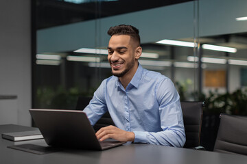 Fototapeta premium A cheerful young middle eastern manager sits at a worktable in a modern office, focused on his notebook as he sends emails and conducts marketing research. The office has a relaxed atmosphere.