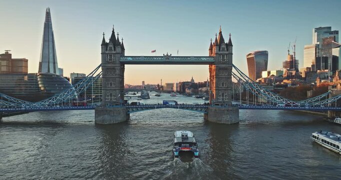 Iconic Tower Bridge at sunset, intricate architecture over River Thames, The Shard, Gherkin, tourist tour boats and Walkie Talkie buildings shaping London cityscape under an evening sky. Drone flight