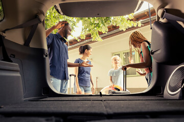 Happy mom with little girl and friends packing the car trunk with luggage and beach essentials for a summer vacation. Diverse group of people prepare for a fun road trip with laughter and joy.